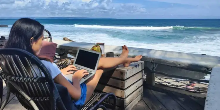 Mujer trabajando en laptop desde una silla de mimbre frente al océano. Sus piernas están apoyadas en una estructura de madera. Cielo azul con nubes.