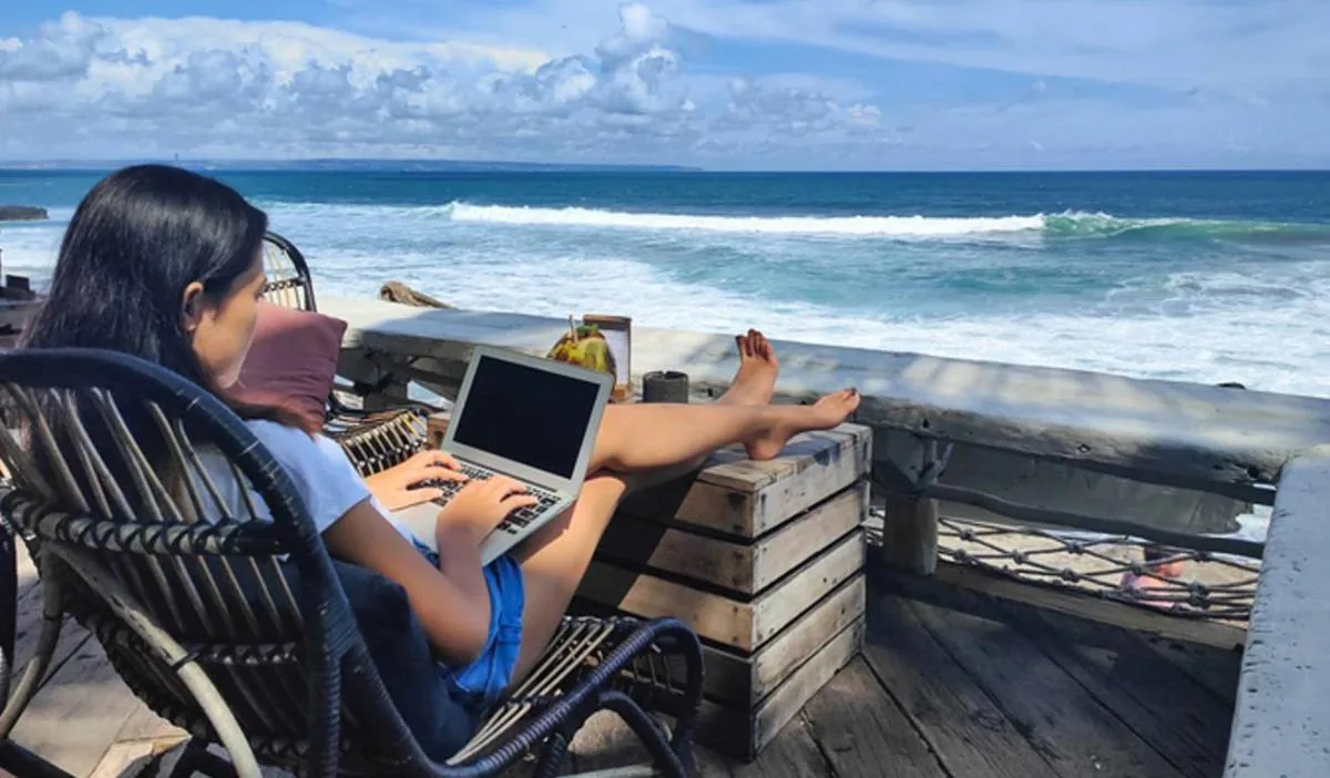 Mujer trabajando en laptop desde una silla de mimbre frente al océano. Sus piernas están apoyadas en una estructura de madera. Cielo azul con nubes.