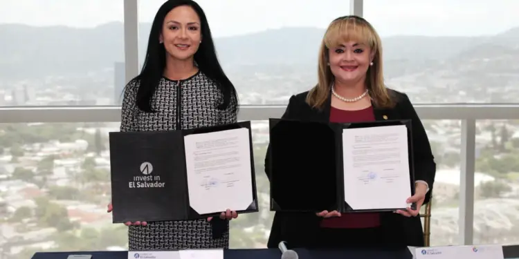 Dos mujeres profesionales sonrientes sostienen documentos firmados en un evento de negocios. Una carpeta muestra 'invest in El Salvador'.