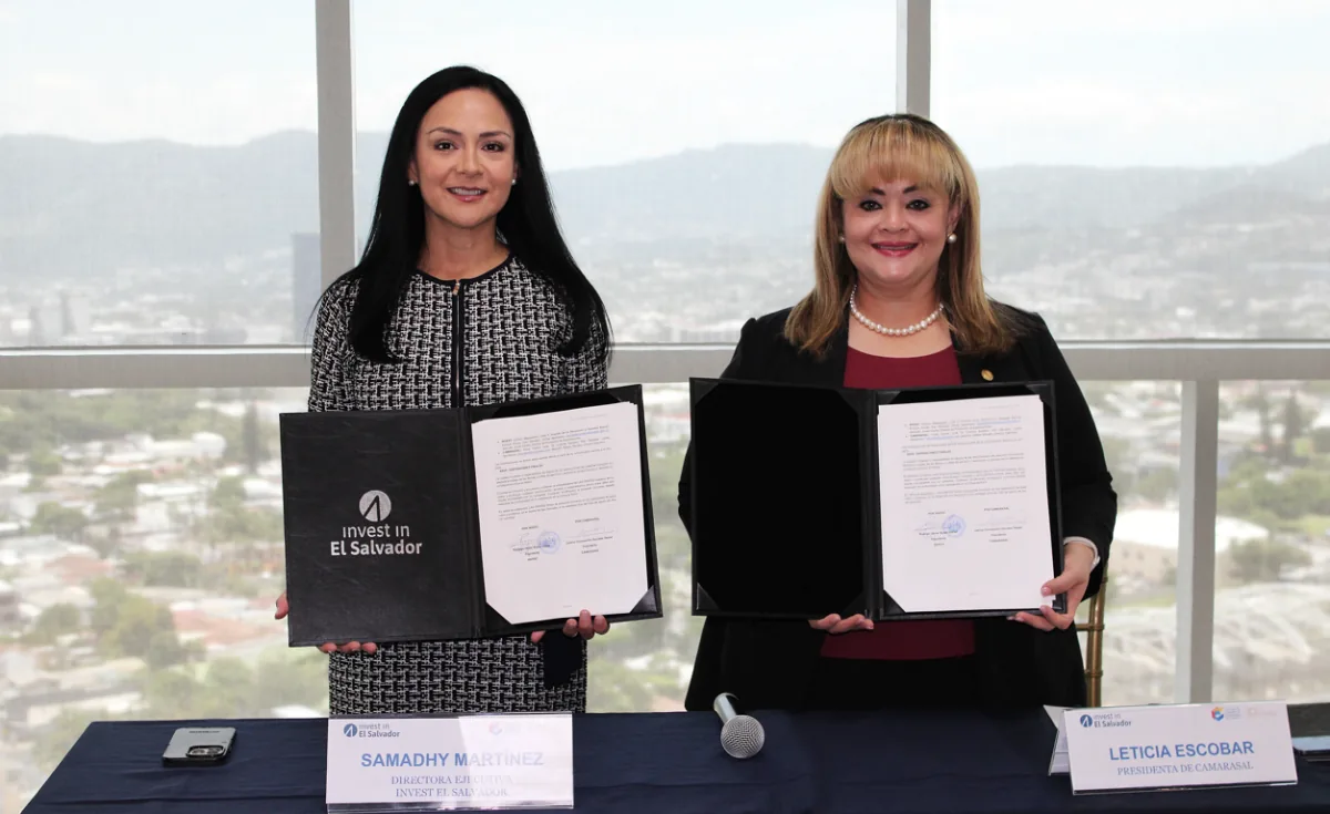 Dos mujeres profesionales sonrientes sostienen documentos firmados en un evento de negocios. Una carpeta muestra 'invest in El Salvador'.