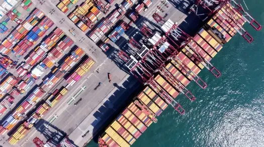 Vista aérea de un puerto comercial con un gran buque de carga atracado, grúas portuarias y miles de contenedores de envío apilados en el muelle.