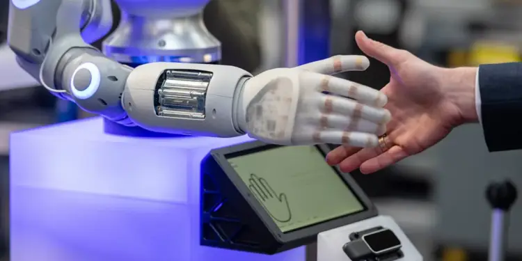 Primer plano de un robot y un humano dándose la mano. El brazo robótico es gris y blanco con luces azules, y la mano es translúcida. La mano humana lleva un anillo.
