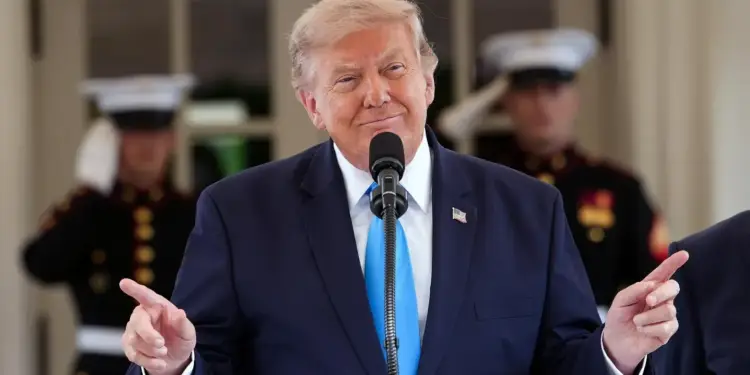 Donald Trump con traje azul y corbata celeste hablando ante un micrófono en una conferencia con guardias de fondo.