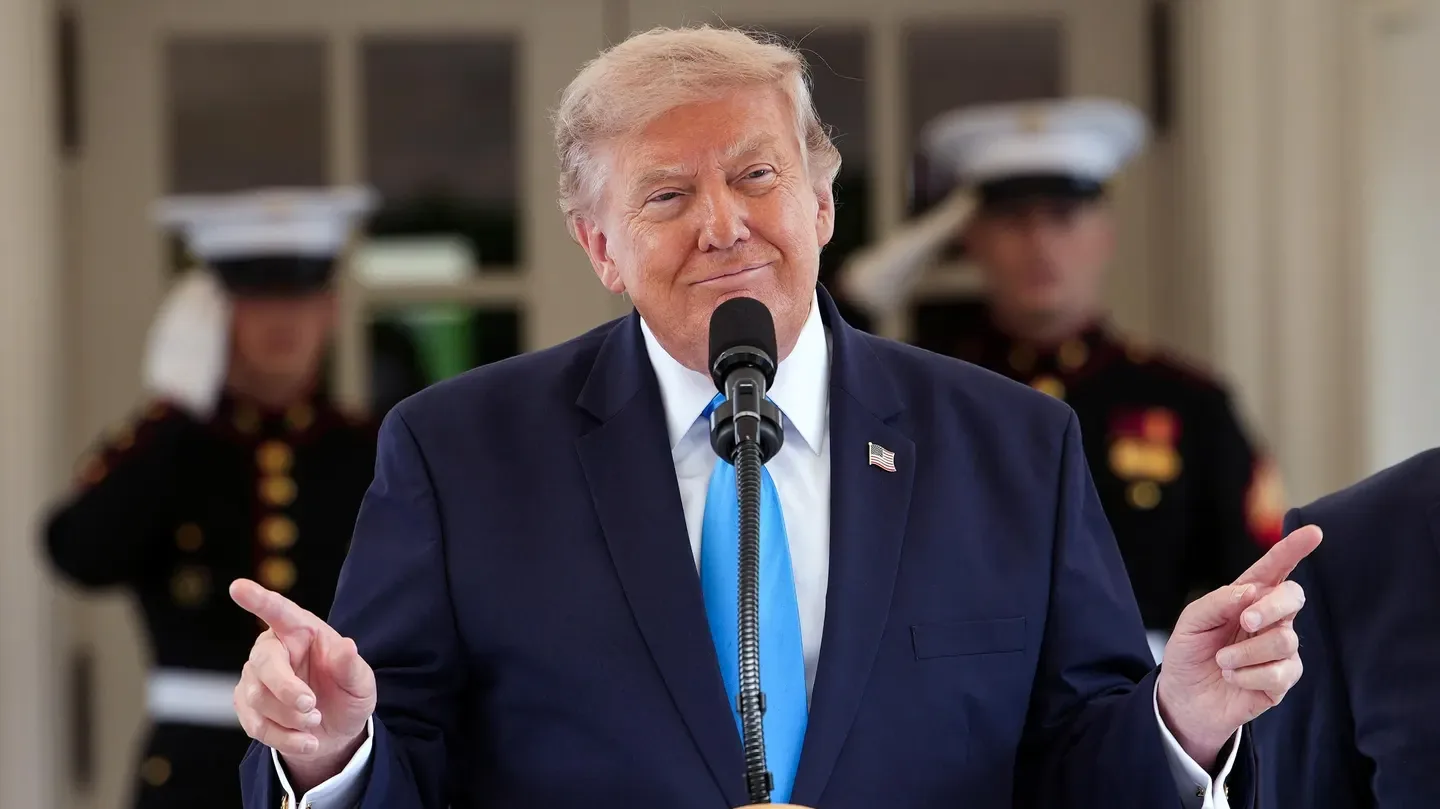 Donald Trump con traje azul y corbata celeste hablando ante un micrófono en una conferencia con guardias de fondo.