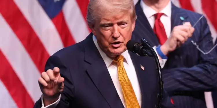 Hombre con traje oscuro y corbata amarilla hablando ante un micrófono frente a banderas de Estados Unidos.
