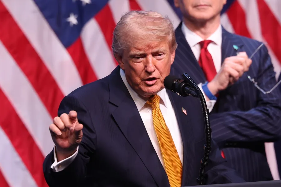 Hombre con traje oscuro y corbata amarilla hablando ante un micrófono frente a banderas de Estados Unidos.