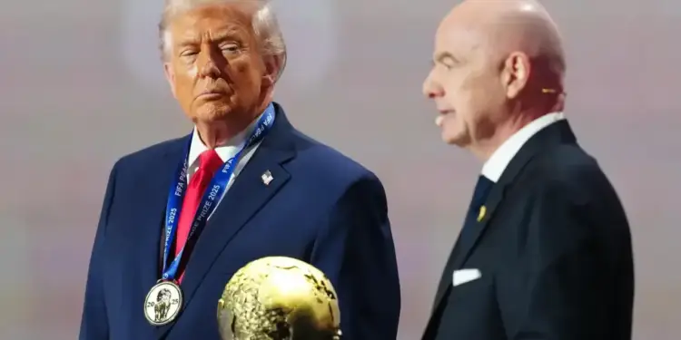 Dos hombres con traje formal en un escenario, uno de ellos con una medalla al cuello y un trofeo dorado en primer plano.