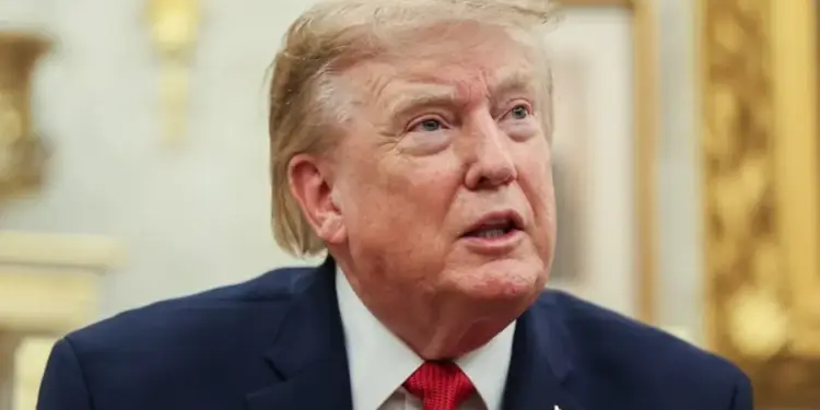 Primer plano de un hombre de mediana edad con cabello rubio, traje azul marino, camisa blanca y corbata roja, con un pin de bandera en la solapa, en un entorno formal.