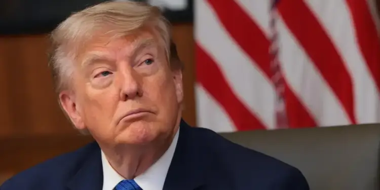Retrato de Donald Trump con traje azul y corbata azul frente a una bandera de Estados Unidos.