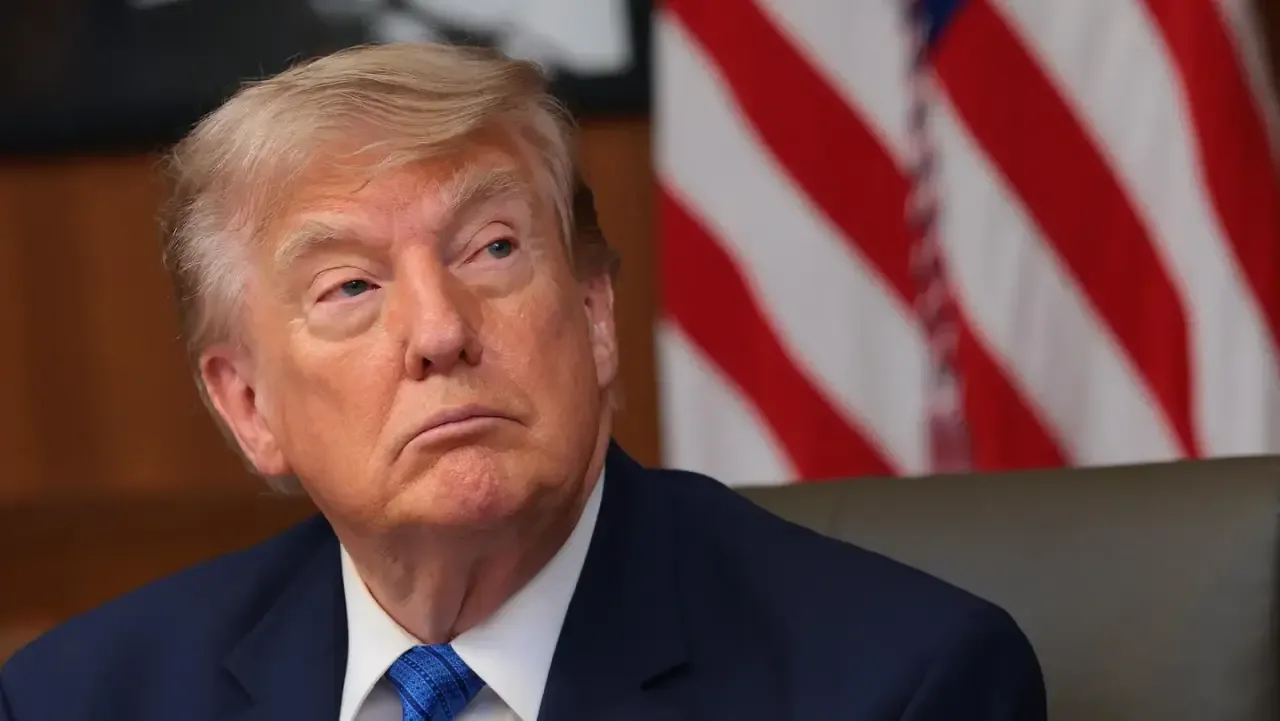 Retrato de Donald Trump con traje azul y corbata azul frente a una bandera de Estados Unidos.