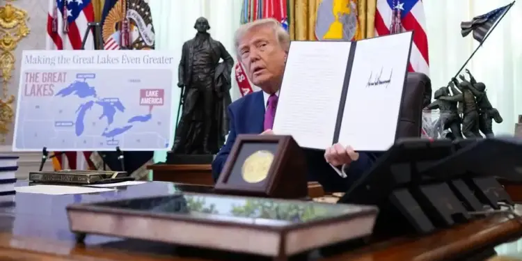 Un hombre con traje y corbata muestra un documento firmado en un despacho oficial con banderas de fondo.