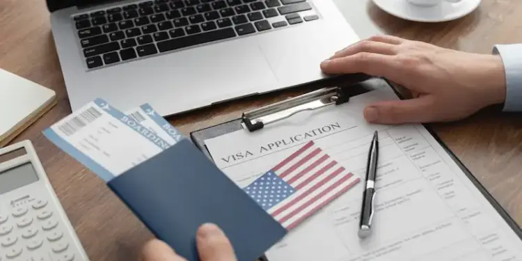 Manos sosteniendo pasaporte con tarjetas de embarque sobre un formulario de solicitud de visa y una laptop.