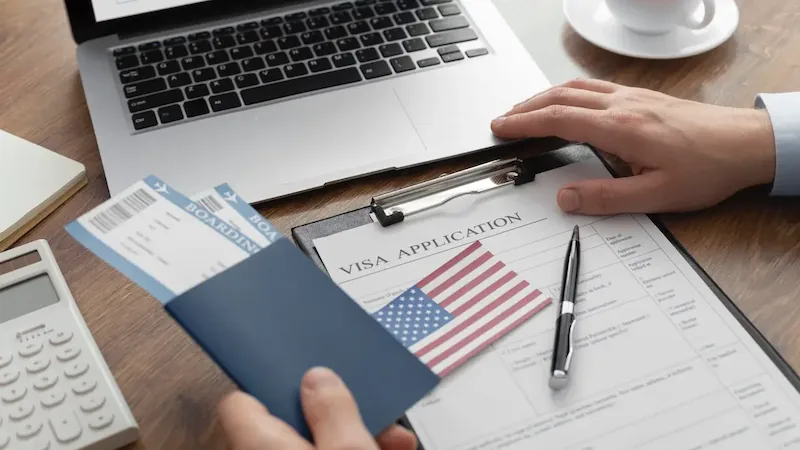 Manos sosteniendo pasaporte con tarjetas de embarque sobre un formulario de solicitud de visa y una laptop.