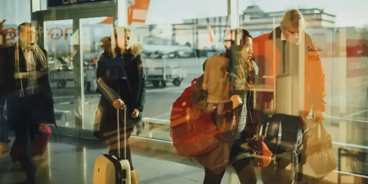 Personas con equipaje en un aeropuerto, vistas a través de un cristal con reflejos. Se observan viajeros y un avión en el exterior.