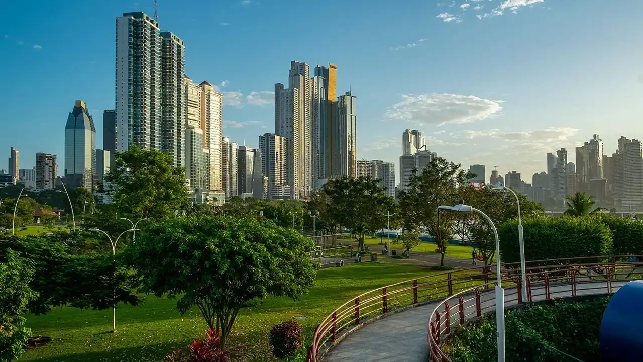 Paisaje urbano con rascacielos altos al fondo y un parque verde con senderos y árboles en primer plano bajo un cielo azul