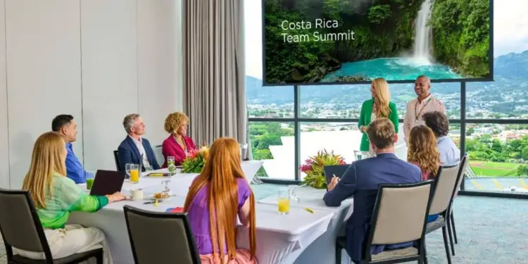 Grupo de profesionales en una sala de juntas corporativa viendo una presentación proyectada sobre una cumbre de equipos.