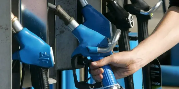Una mano sostiene una manguera de combustible azul en una estación de servicio. Se observan otros surtidores de gasolina en el fondo, algunos azules y otros negros, indicando el acto de repostar.