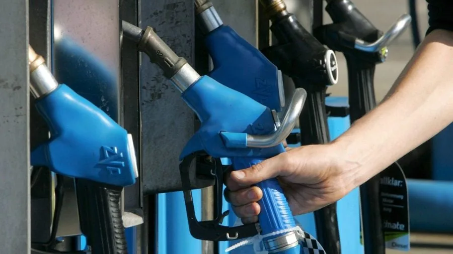 Una mano sostiene una manguera de combustible azul en una estación de servicio. Se observan otros surtidores de gasolina en el fondo, algunos azules y otros negros, indicando el acto de repostar.
