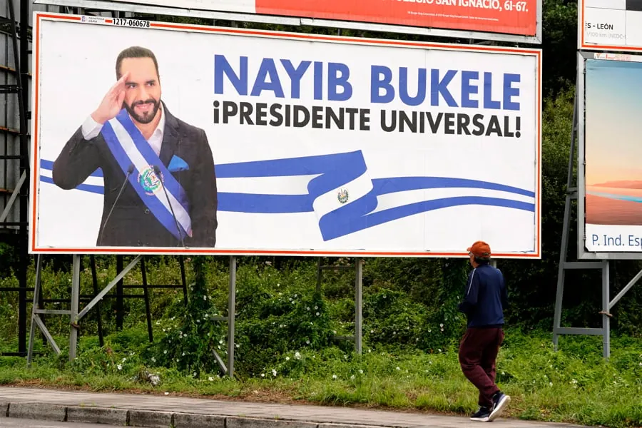Gran valla publicitaria con Nayib Bukele en traje y banda presidencial, saludando. Texto: '¡PRESIDENTE UNIVERSAL!'. Persona camina.