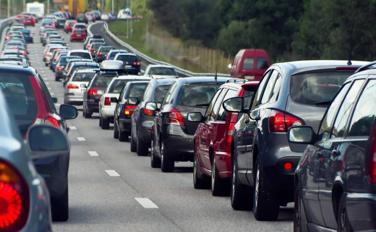 Vista trasera de una carretera con múltiples carriles llena de coches detenidos o moviéndose lentamente, formando un atasco. Se aprecian vehículos de diversos colores en una fila interminable bajo un cielo diurno.