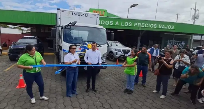 Grupo de personas cortando una cinta azul frente a un camión blanco de carga frente a un establecimiento comercial.