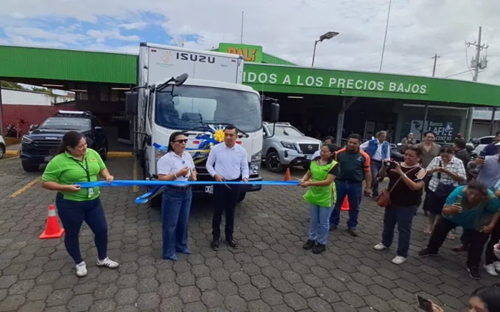 Grupo de personas cortando una cinta azul frente a un camión blanco de carga frente a un establecimiento comercial.