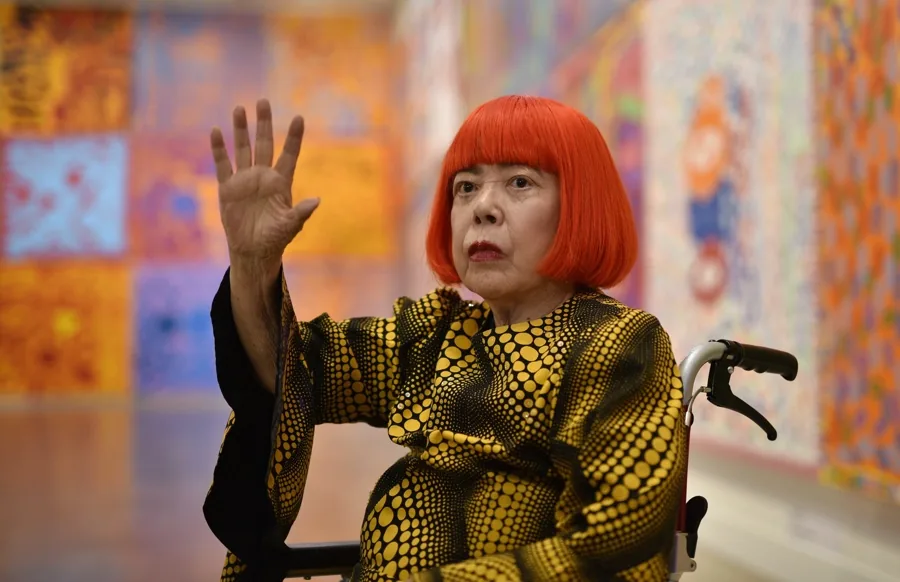 Retrato de persona mayor con cabello rojo y atuendo de lunares, sentada en silla de ruedas, levantando la mano. Fondo de galería de arte abstracto.