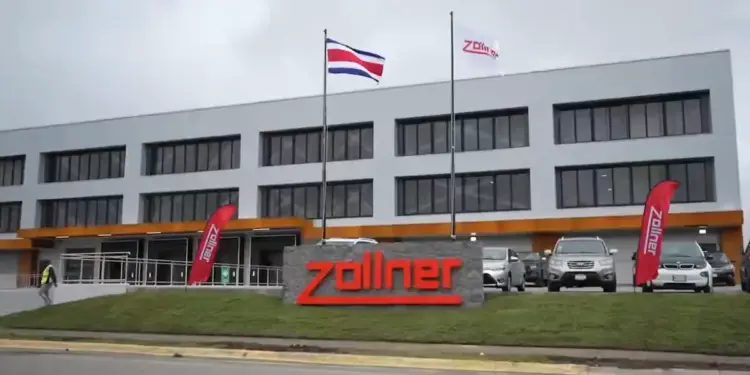 Edificio corporativo moderno de Zollner con fachada gris y naranja, logo rojo en piedra, bandera de Costa Rica y vehículos en el estacionamiento.