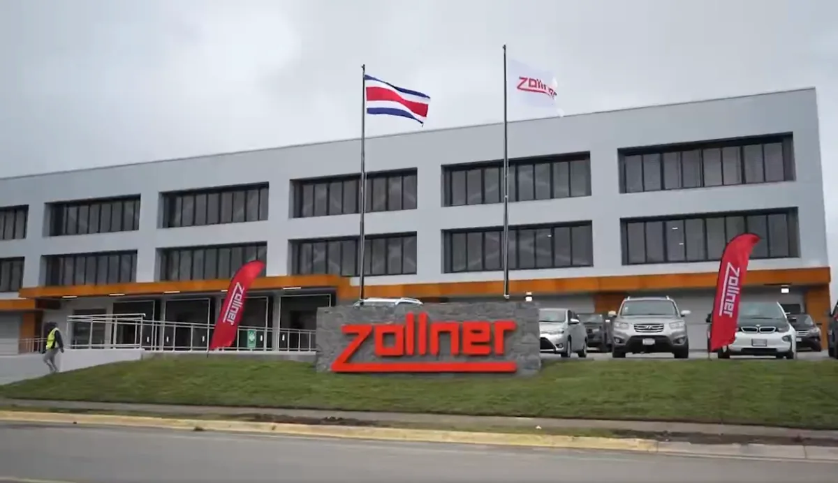 Edificio corporativo moderno de Zollner con fachada gris y naranja, logo rojo en piedra, bandera de Costa Rica y vehículos en el estacionamiento.