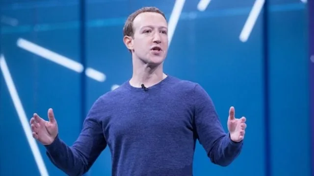 Hombre caucásico de mediana edad con camisa azul oscuro, hablando y gesticulando con ambas manos en un escenario azul con luces diagonales.