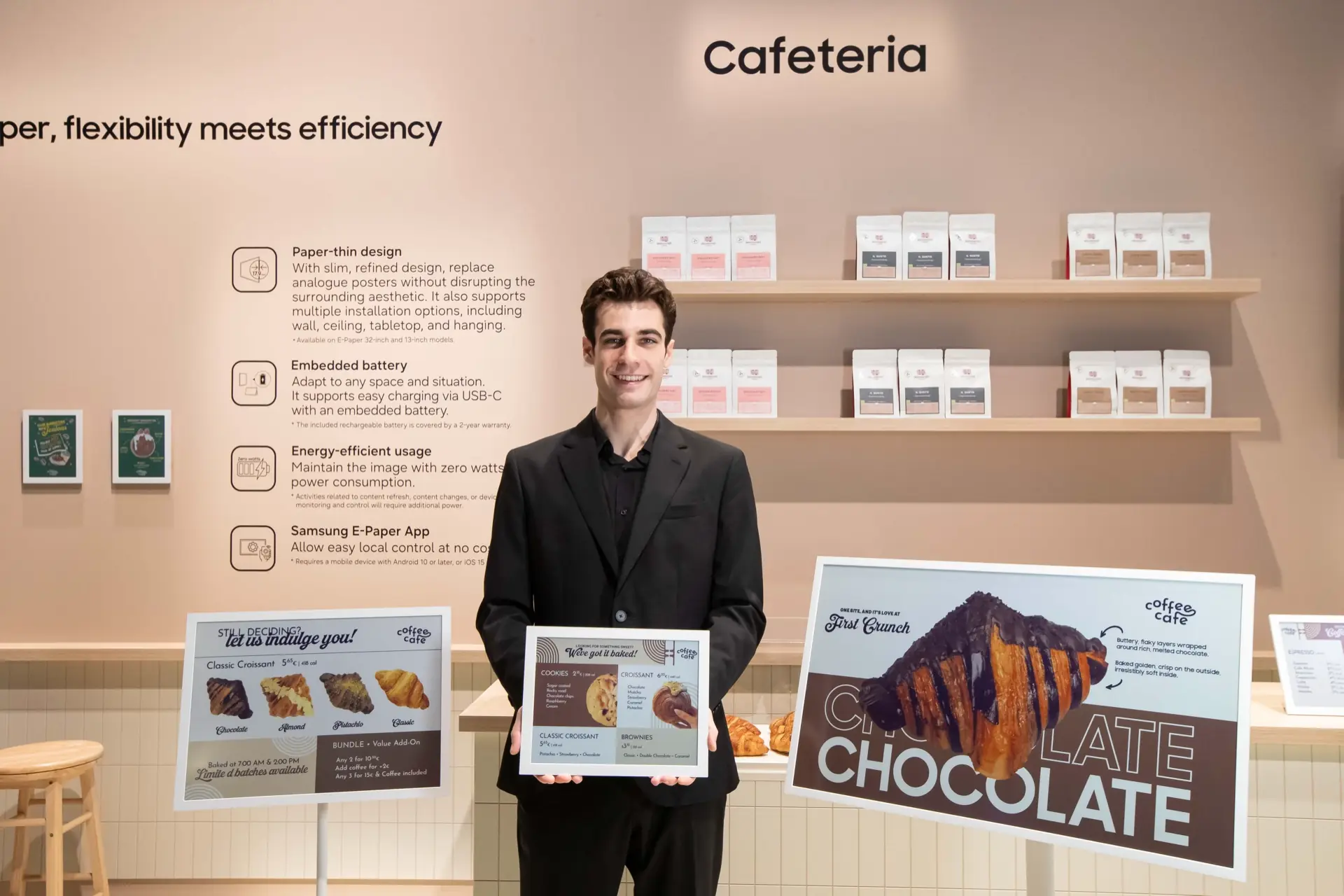 Hombre de traje negro sonriendo mientras sostiene un cartel publicitario digital en una cafetería moderna.