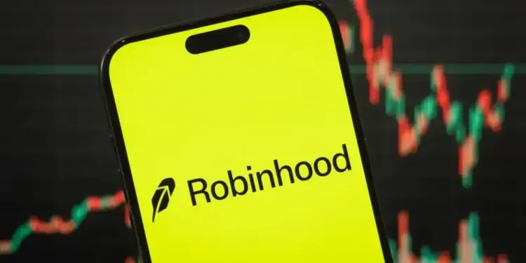 Primer plano de un smartphone con la aplicación Robinhood abierta en pantalla de color amarillo brillante, con un gráfico de velas de bolsa en segundo plano.