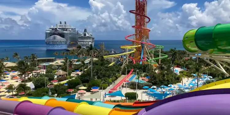 Vista panorámica de un parque acuático tropical con coloridos toboganes y dos grandes cruceros en el mar de fondo.