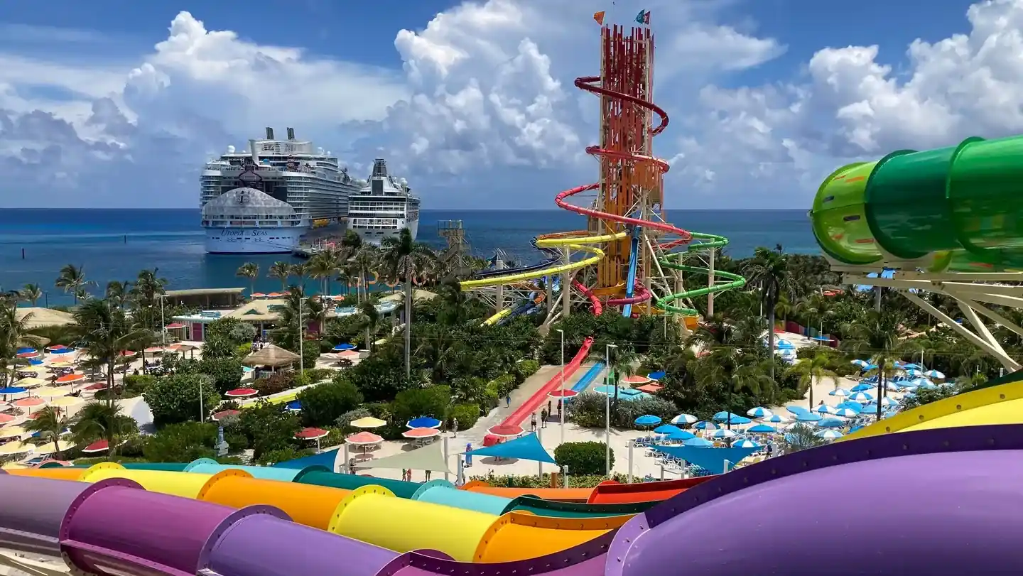 Vista panorámica de un parque acuático tropical con coloridos toboganes y dos grandes cruceros en el mar de fondo.