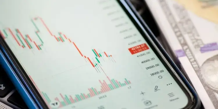 Pantalla de móvil con gráfico de velas financieras (rojas y verdes) y datos numéricos. Se aprecian billetes de dólar y un teclado.