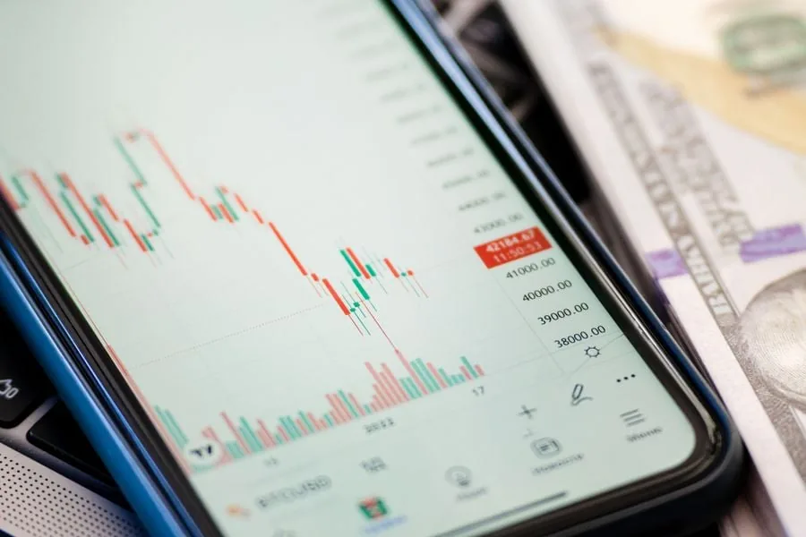 Pantalla de móvil con gráfico de velas financieras (rojas y verdes) y datos numéricos. Se aprecian billetes de dólar y un teclado.