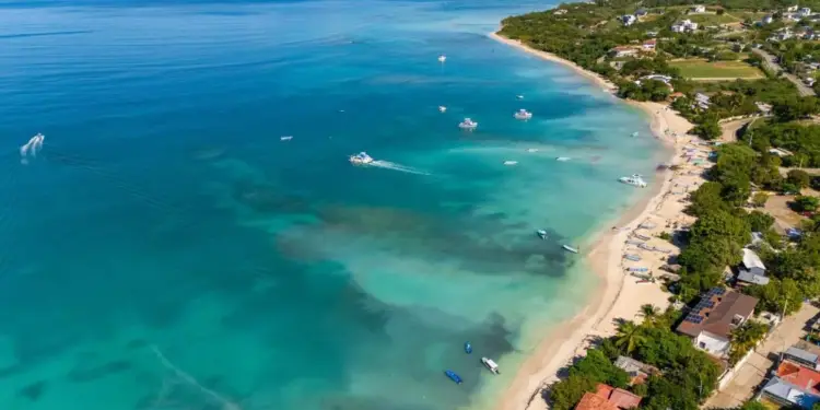 Vista aérea de una costa tropical con aguas turquesas, playa de arena, múltiples embarcaciones y viviendas con paneles solares en la orilla.