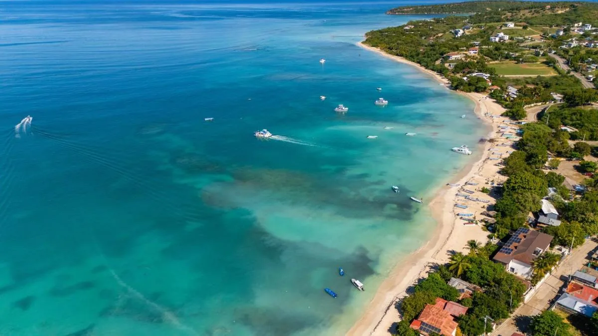 Vista aérea de una costa tropical con aguas turquesas, playa de arena, múltiples embarcaciones y viviendas con paneles solares en la orilla.