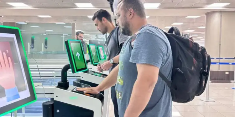 Dos hombres utilizan kioscos de auto-servicio con pantallas verdes y escáneres biométricos. Uno coloca su mano en el lector, agilizando trámites.