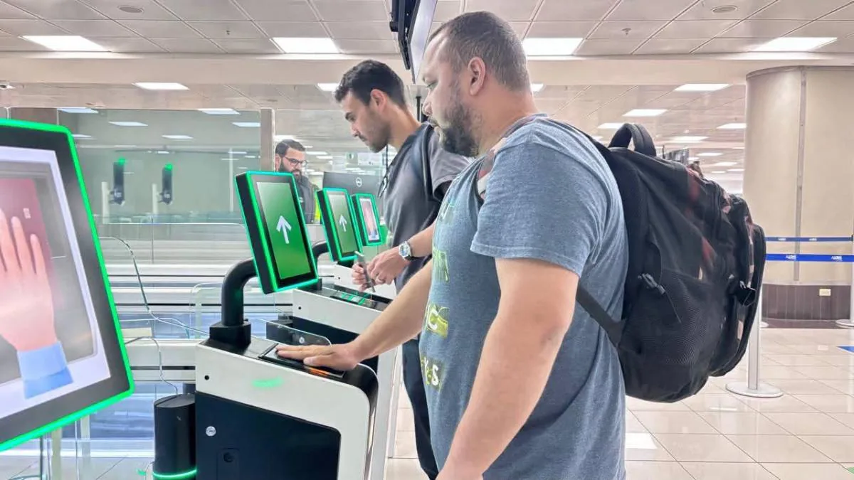 Dos hombres utilizan kioscos de auto-servicio con pantallas verdes y escáneres biométricos. Uno coloca su mano en el lector, agilizando trámites.