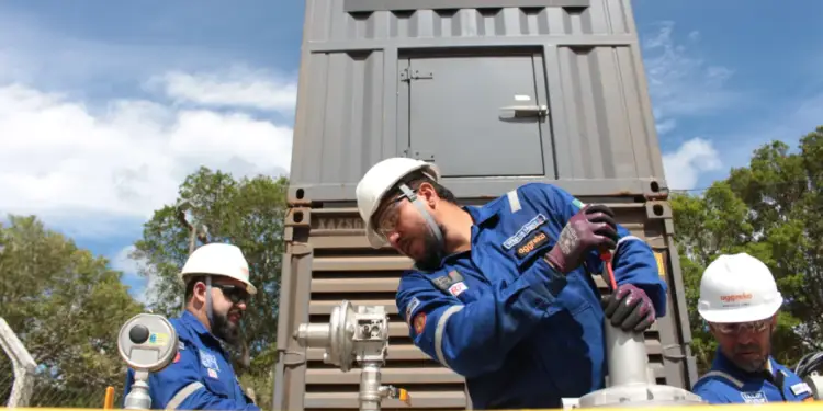 Tres técnicos de Aggreko con uniformes azules y cascos blancos operan equipos industriales bajo un cielo azul. Un contenedor gris con el logo Aggreko es visible.
