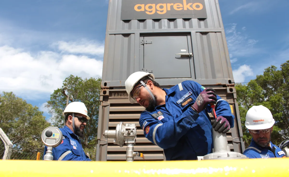 Tres técnicos de Aggreko con uniformes azules y cascos blancos operan equipos industriales bajo un cielo azul. Un contenedor gris con el logo Aggreko es visible.