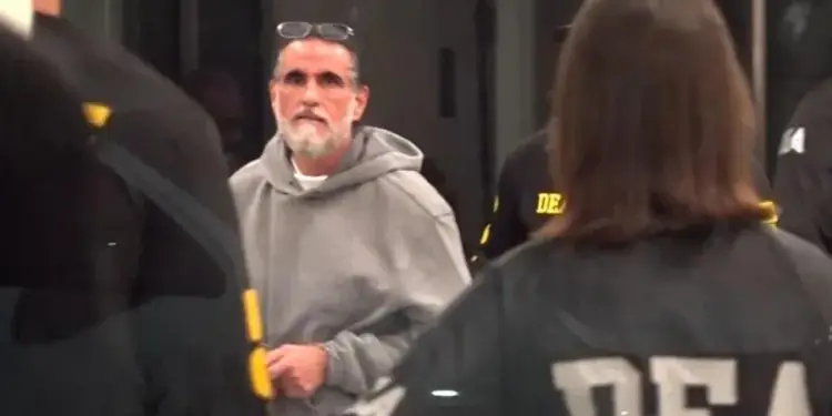 Un hombre con barba y sudadera gris rodeado por personas con uniformes con la inscripción DEA en la espalda.
