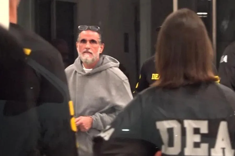 Un hombre con barba y sudadera gris rodeado por personas con uniformes con la inscripción DEA en la espalda.