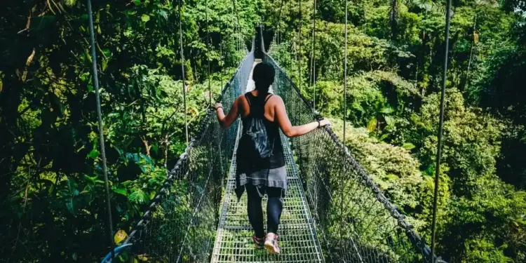 Una persona con mochila caminando por un puente colgante de metal rodeado de vegetación densa.