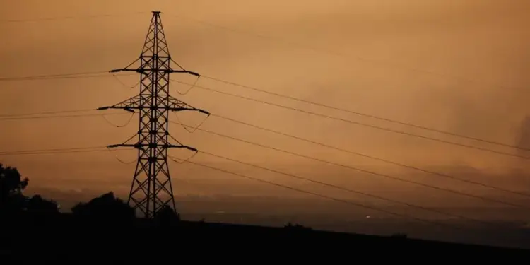 Silueta de una torre de alta tensión con cables eléctricos sobre un fondo de cielo naranja al atardecer.