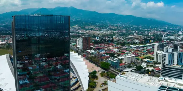 Vista aérea de un distrito financiero con modernos rascacielos de vidrio y montañas al fondo bajo un cielo nublado.