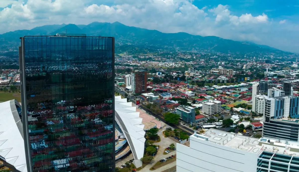Vista aérea de un distrito financiero con modernos rascacielos de vidrio y montañas al fondo bajo un cielo nublado.