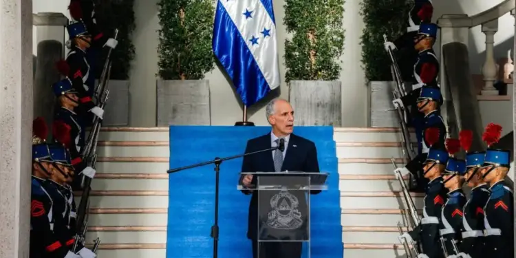 Hombre de traje hablando en un atril frente a una bandera y una guardia de honor militar.