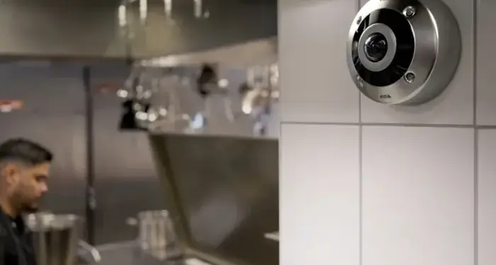 Cámara de vigilancia redonda instalada en una pared de azulejos blancos dentro de una cocina profesional con un trabajador al fondo.